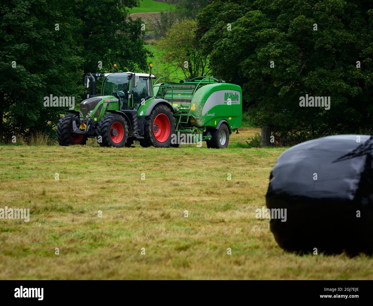 Hay or silage making (farmer in farm tractor at work in field, pulling baler, collecting dry grass & round black bale wrapped - Yorkshire England, UK. Stock Photo
