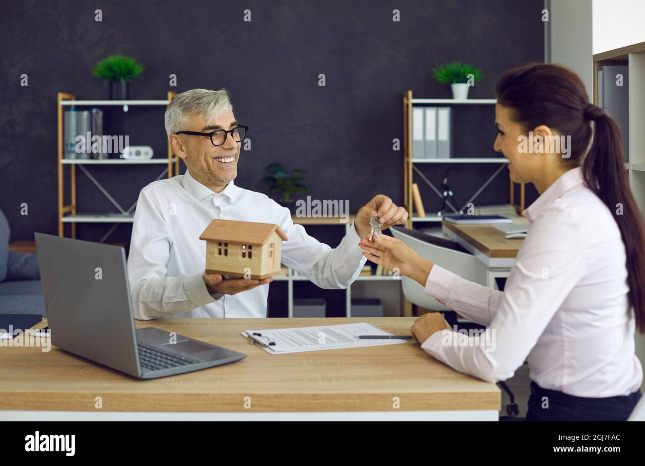 Male real estate agent in office meeting hands over keys to his new home to happy young woman. Stock Photo