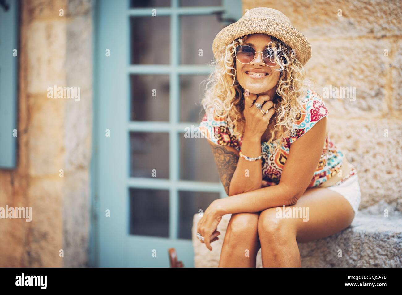 Portrait of beautiful smiling hipster young woman in sunglasses and straw hat sitting outdoors on a bright sunny day. Stylish tattooed woman in good m Stock Photo