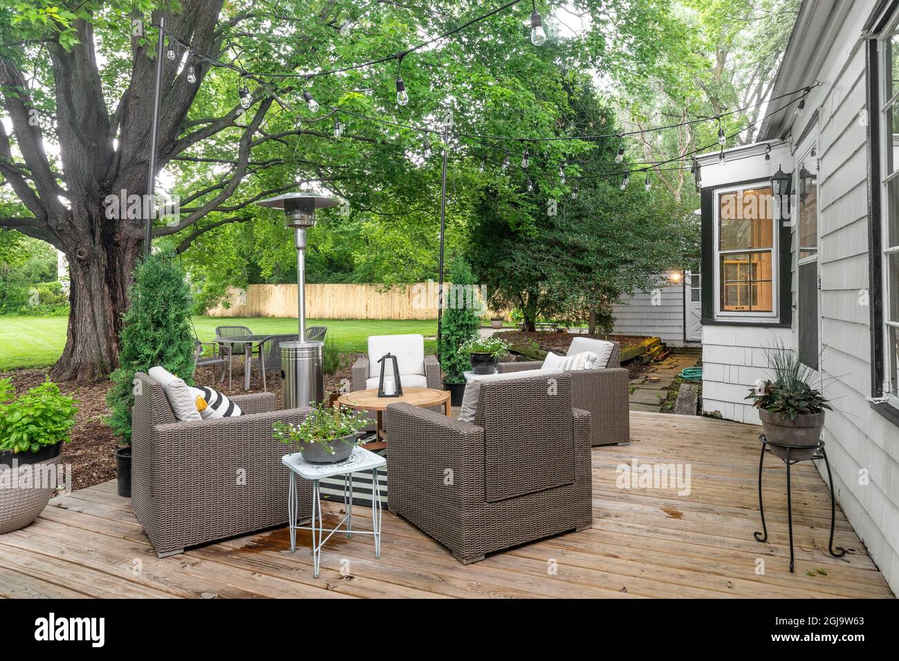 A cozy backyard patio of a white farmhouse with furniture, string lights, and wooden deck. Stock Photo
