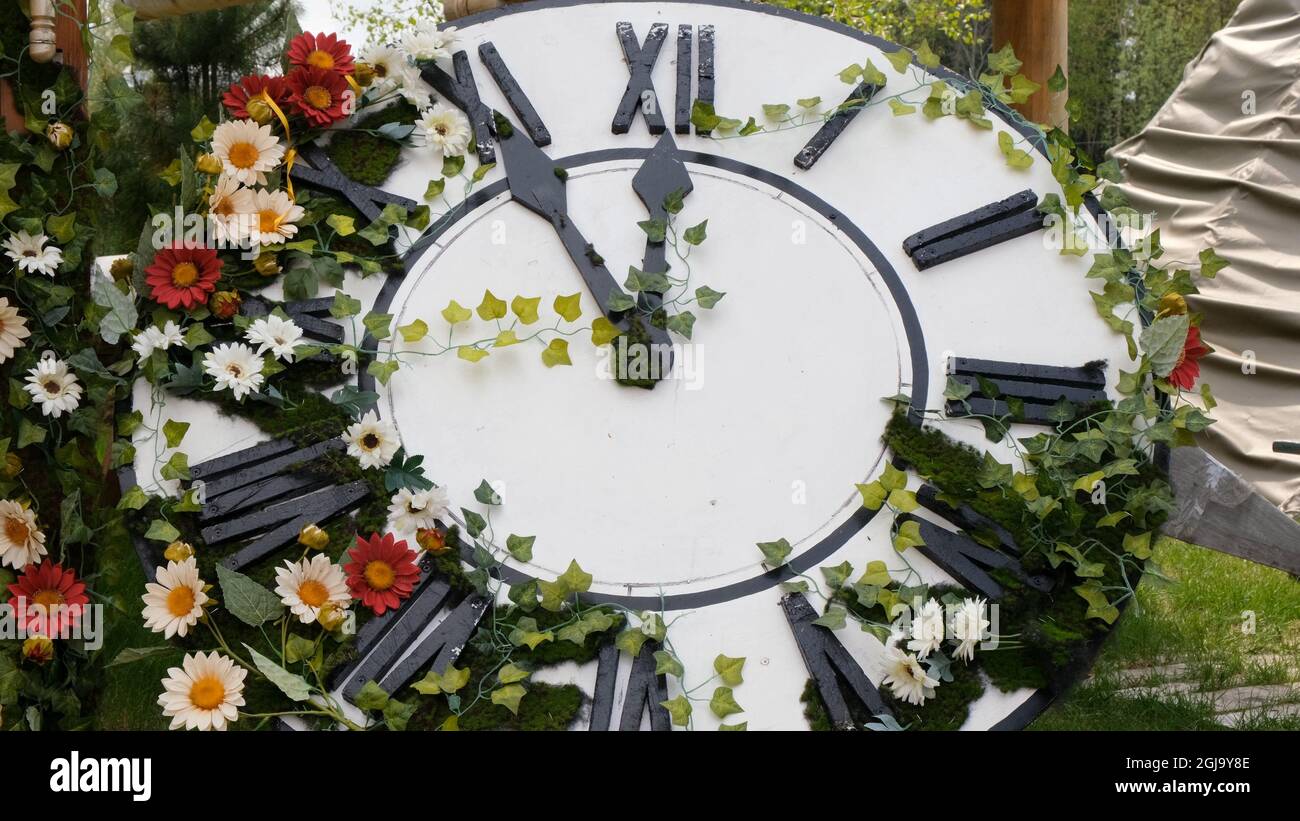 Large wall clock on the field in blooming flowers, showing the time 11: ...