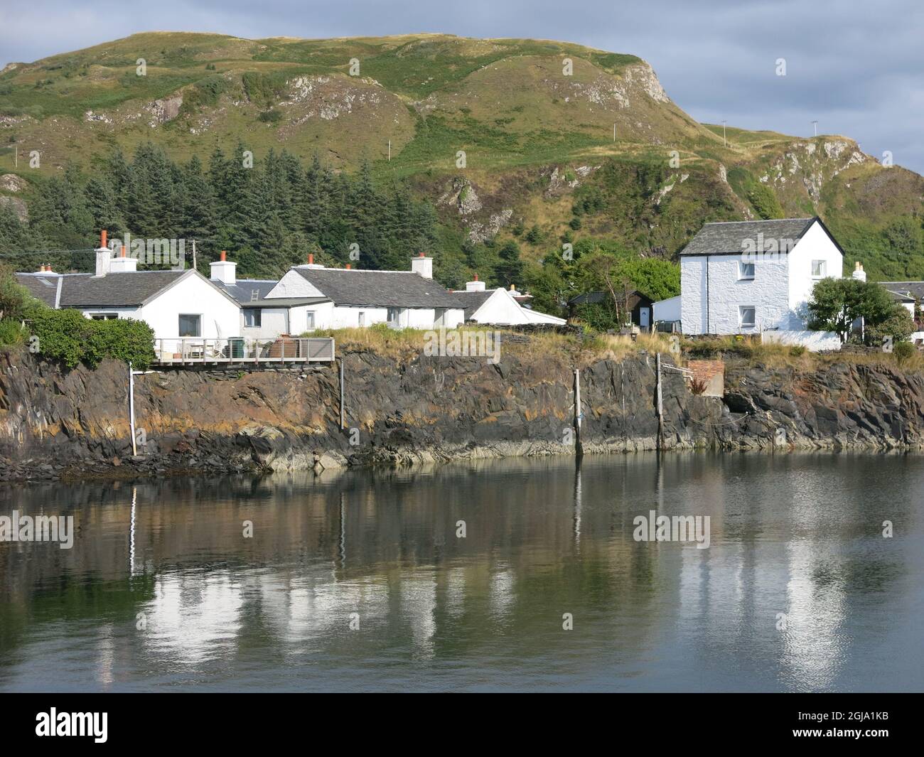 The picturesque village of Ellenabeich with traditional white-washed cottages set against a craggy mountain backdrop; summer 2021, the Inner Hebrides. Stock Photo