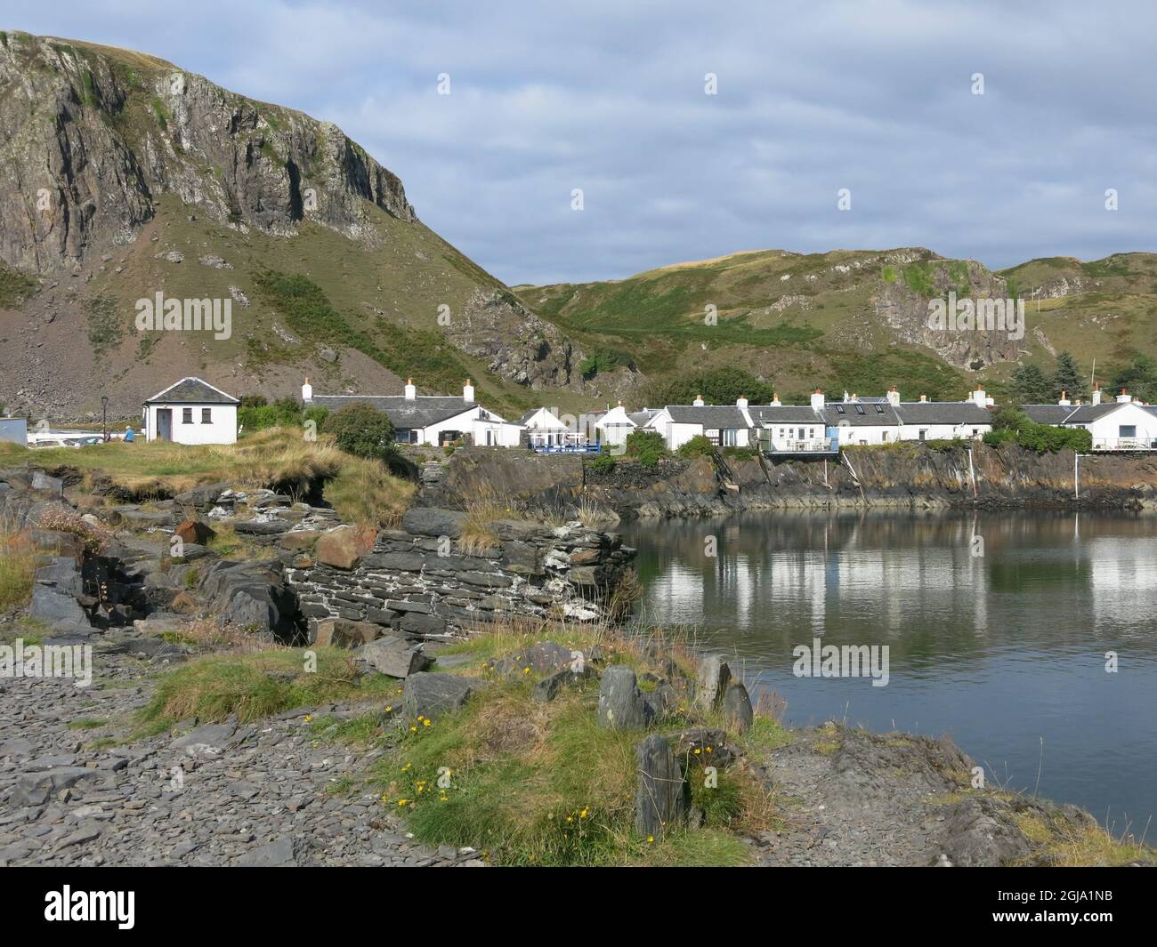 The picturesque village of Ellenabeich with traditional white-washed cottages set against a craggy mountain backdrop; summer 2021, the Inner Hebrides. Stock Photo