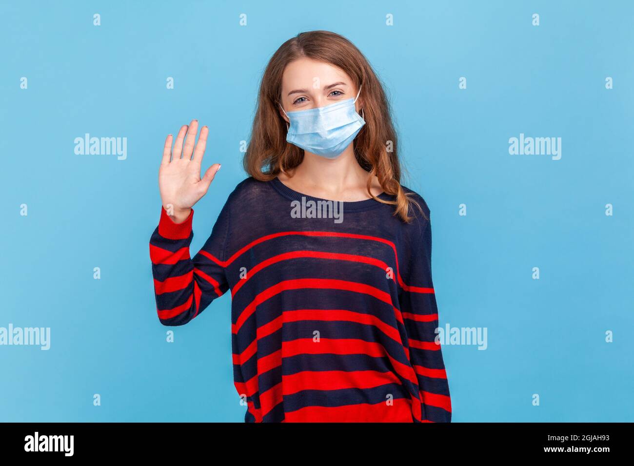 Portrait of happy excited woman wearing striped casual sweater and surgical medical mask standing, looking at camera and waving her hand to greeting, Stock Photo