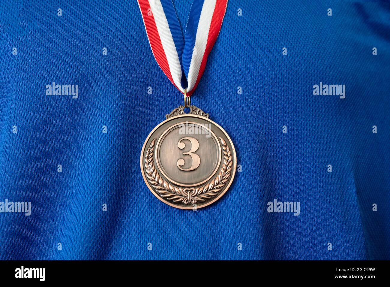 Medal bronze. Winner prize award hanging with red blue color ribbon on athlete chest. Trophy in sport for third place champion on blue color shirt bac Stock Photo