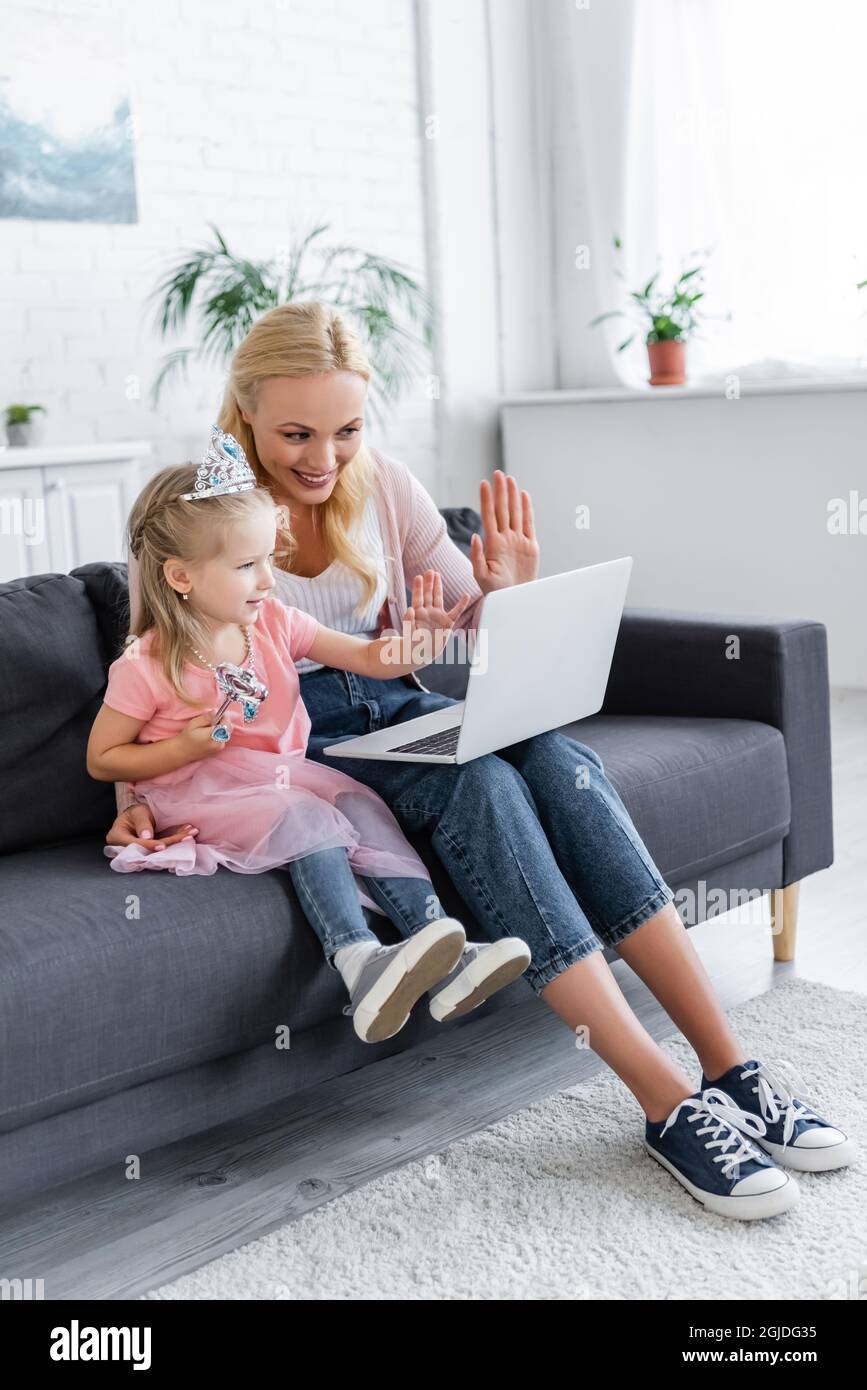 cheerful woman and girl in toy crown waving hands during video call on laptop Stock Photo