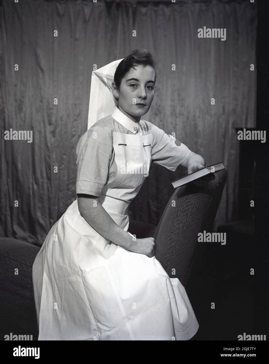 1955,  historical, a nurse in her uniform of this era sitting posing for her photo holding a small book , England, UK. Stock Photo