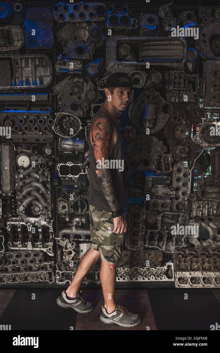 Young handsome caucasian man with a sleeve tattoo wearing military shorts and cap walking by the wall with iron mechanisms.  Stock Photo