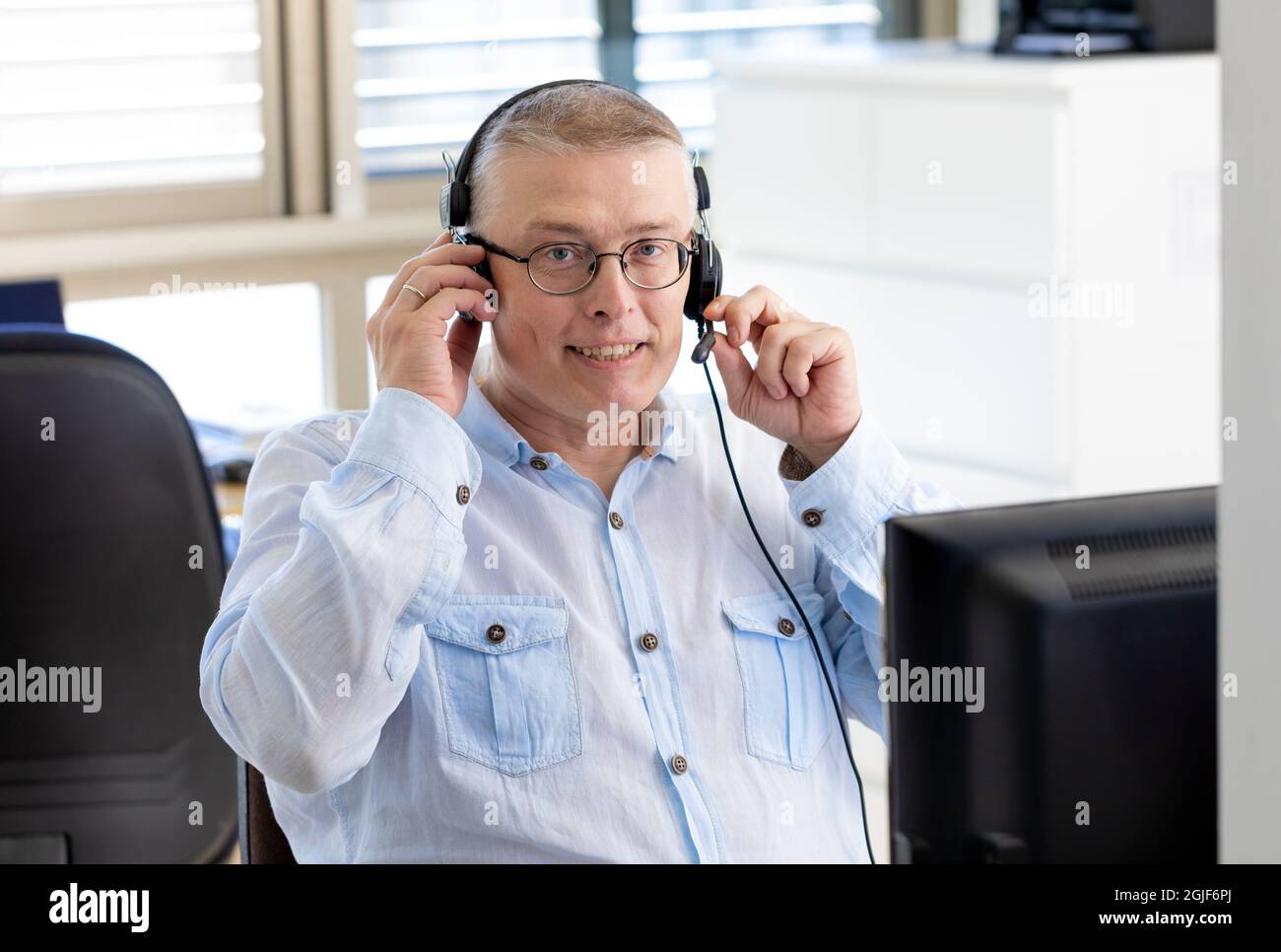 a man works as a manager in an office and talks with clients on the internet through a computer with headphones with a microphone. man of European appearance in a blue shirt sits in the office with his back to the window.  Stock Photo