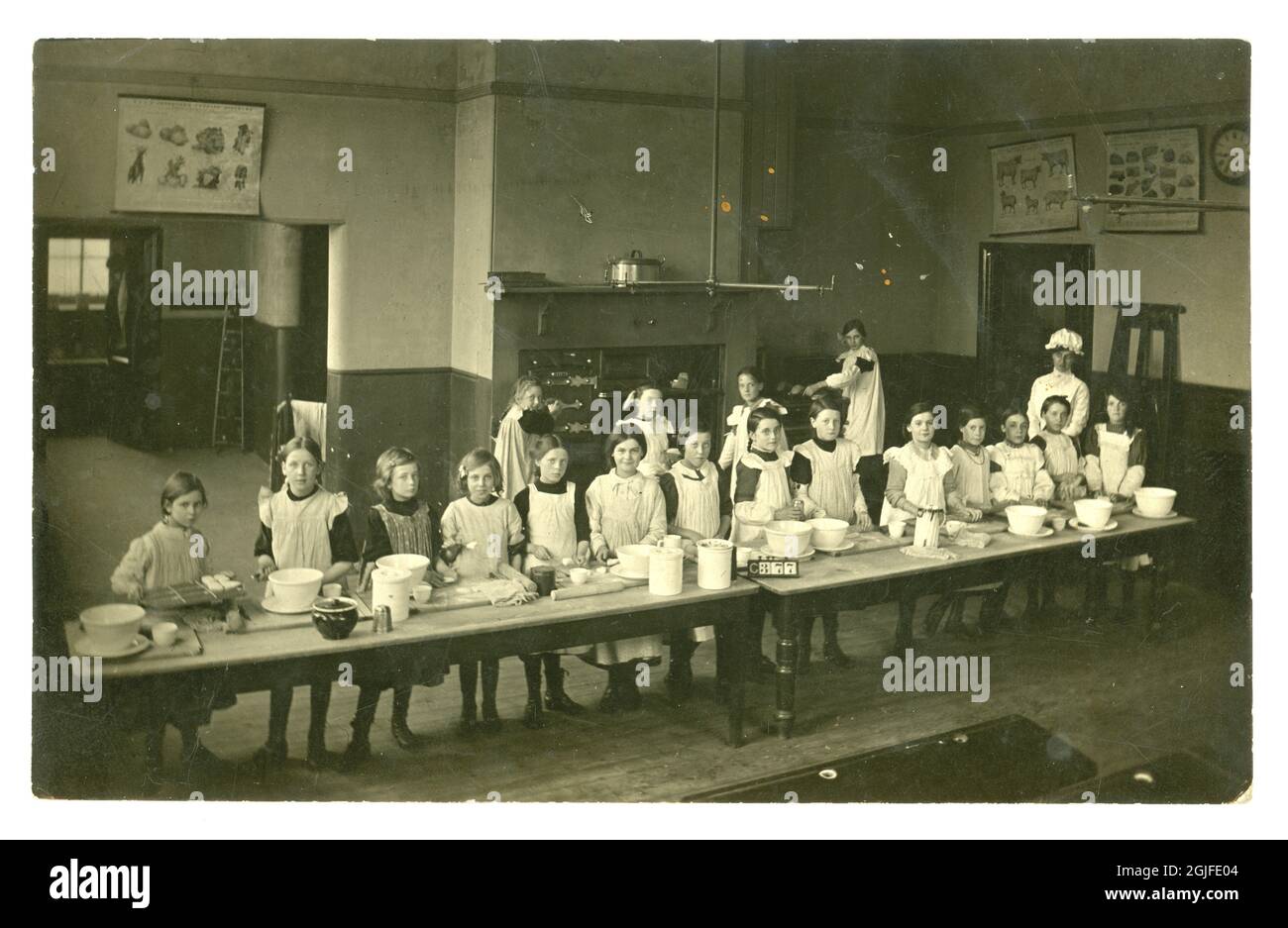Original early 1900's postcard, girls in cookery classroom working at long tables, vegetable, meat cuts and livestock charts on the walls, amazing oven range, baking bread and scones - London, England, U.K. circa 1903 Stock Photo