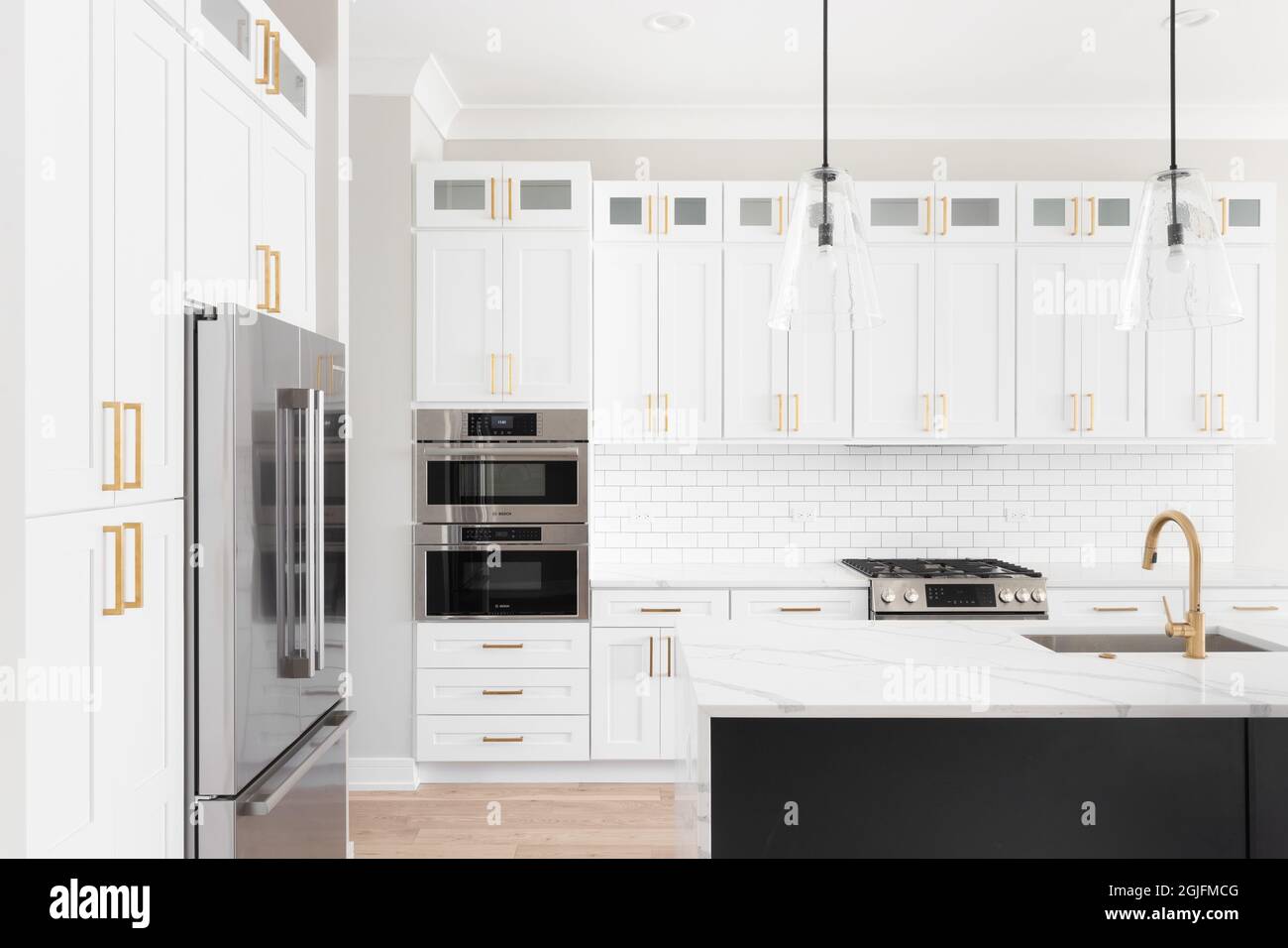 A luxury white kitchen with black pendant lights hanging above a waterfall granite island, stainless steel Bosch appliances, and gold hardware. Stock Photo