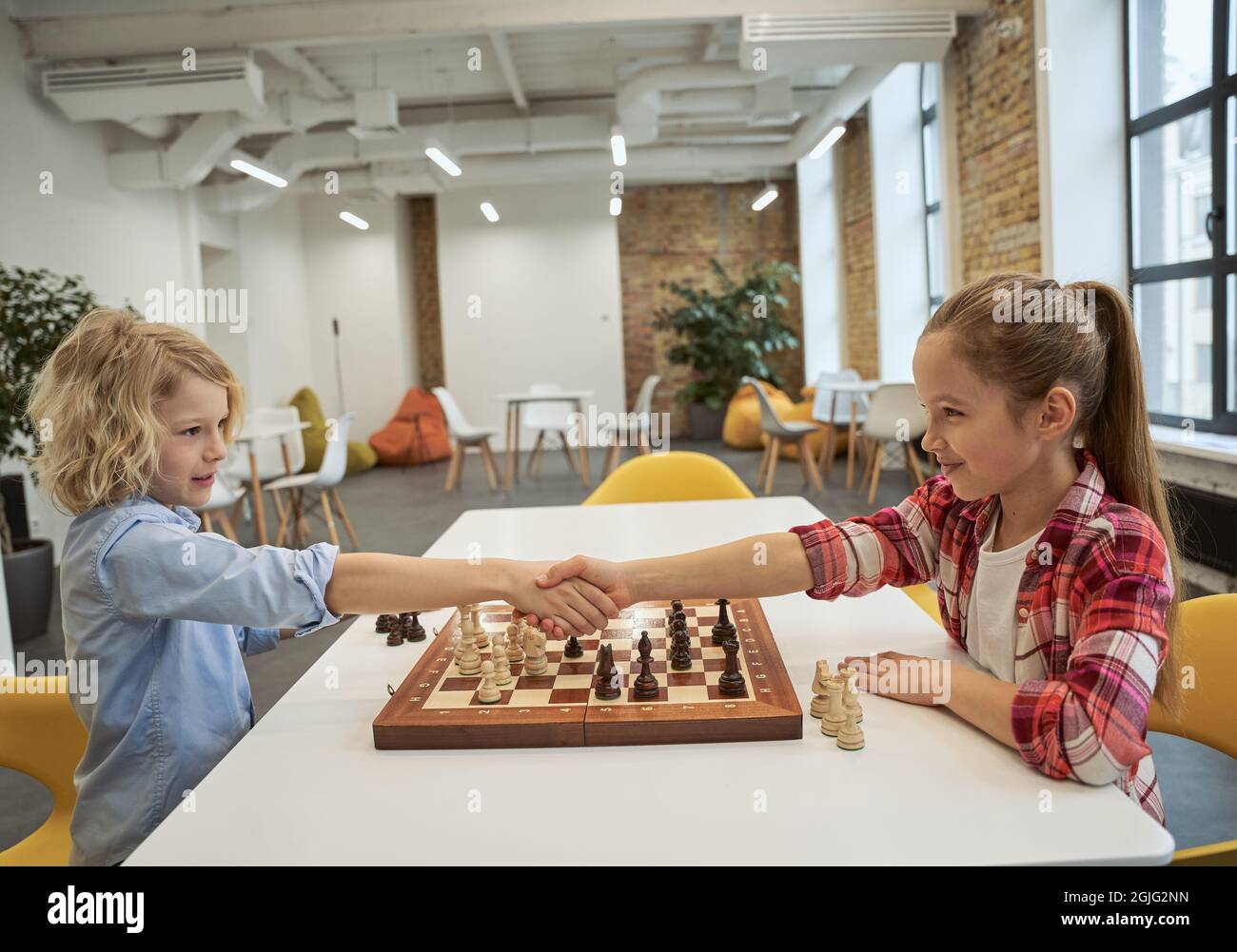 Thank you. Caucasian boy and girl looking at each other and shaking hands after playing chess, sitting together at the table in school Stock Photo