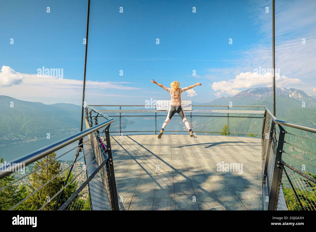 Woman jumping on Aussichtsplattform platform of Cardada-Cimetta mount in Switzerland. Skyline of Locarno on Cardada mount. Lake Langensee cityscape in Stock Photo