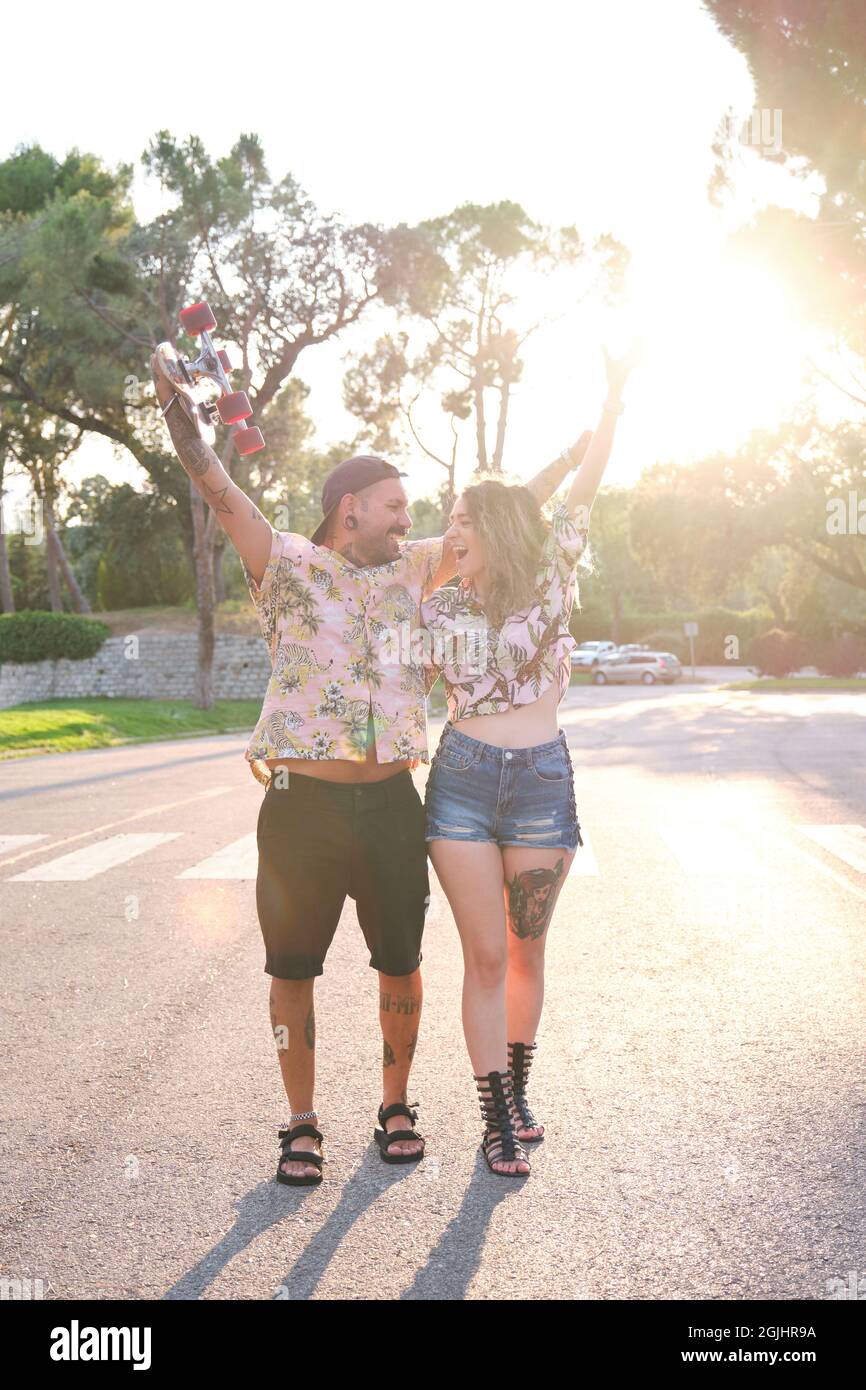 Young cool tattooed couple shout, rise their arms and laugh with a skateboard. Stock Photo