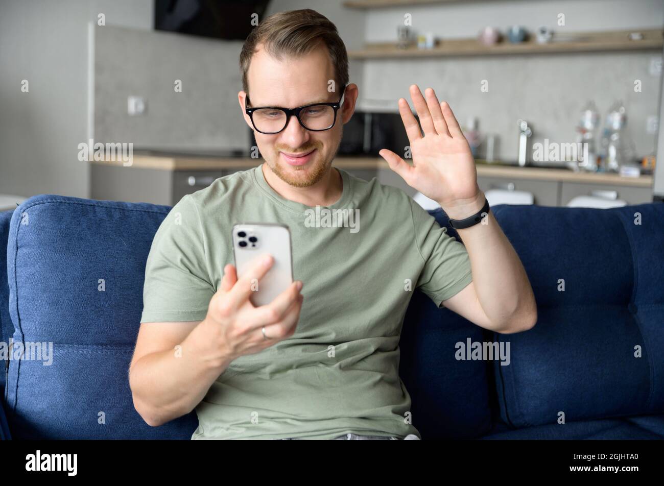 A young handsome guy using a smartphone for video call, smiling man in casual wear is waving hello into webcam, has virtual meeting with friends or employees sitting on the couch at home Stock Photo