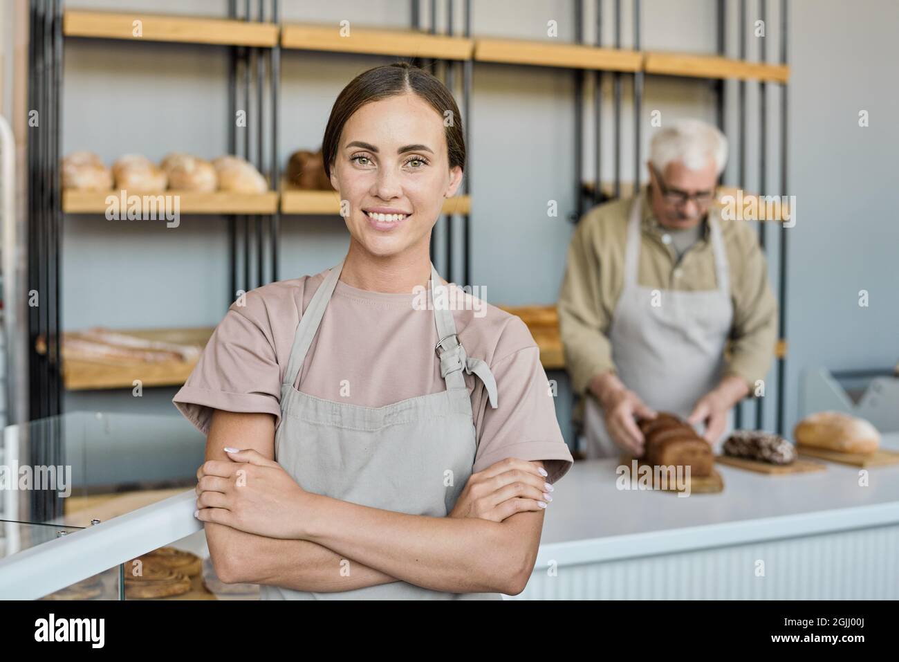 Portrait of happy young brunette woman in apron standing with crossed arms in bakery shop owned by her father Stock Photo