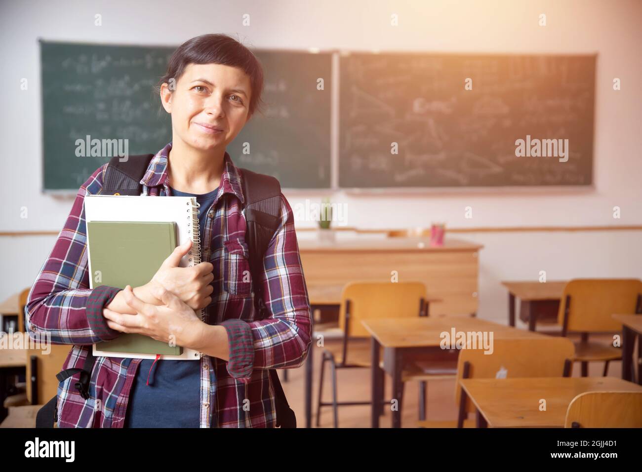 Smiling student girl wearing school backpack and holding exercise book in classroom Stock Photo