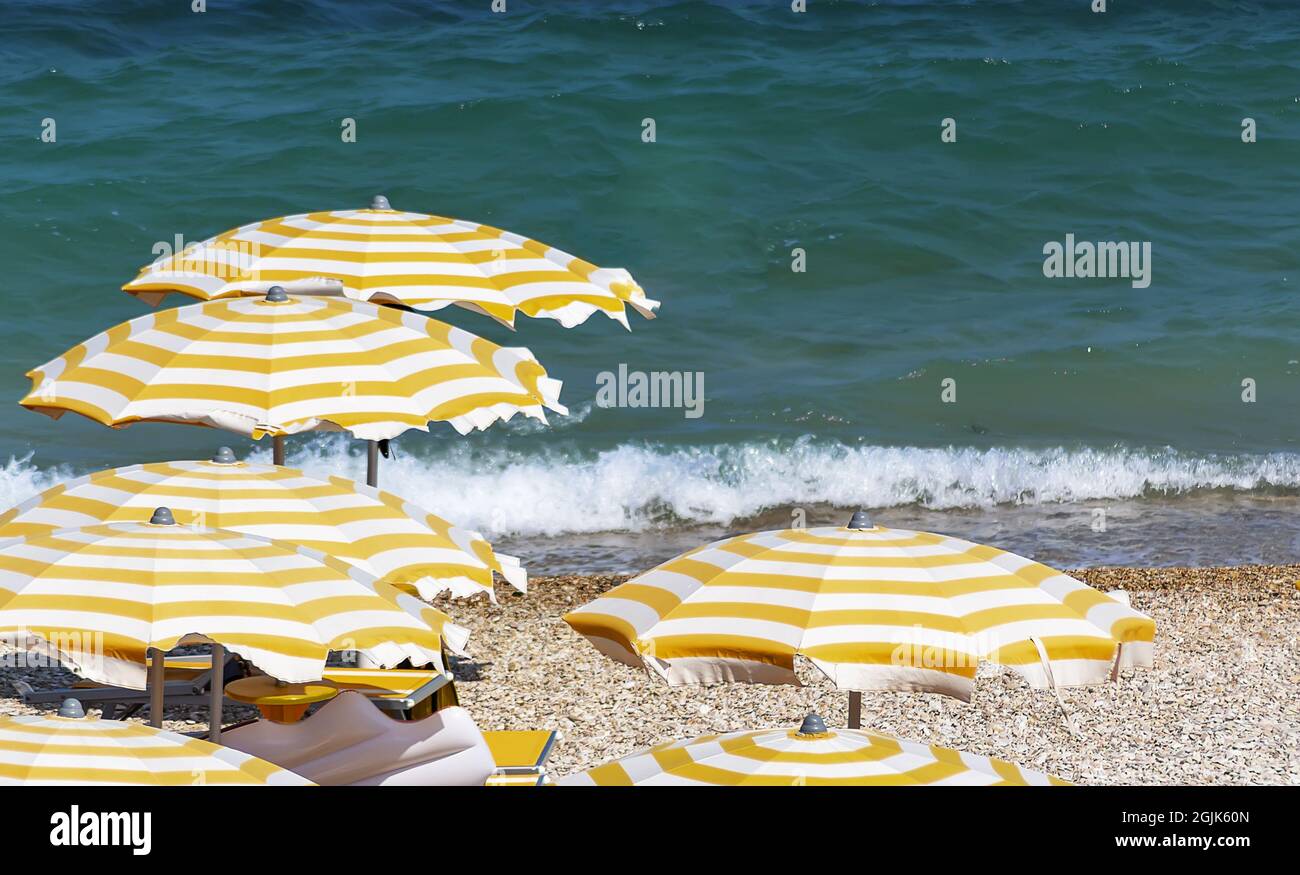 Yellow and white striped umbrellas open in a pebbly beach during a windy summer day. Sea waves on the background. Holidays and relaxation Stock Photo