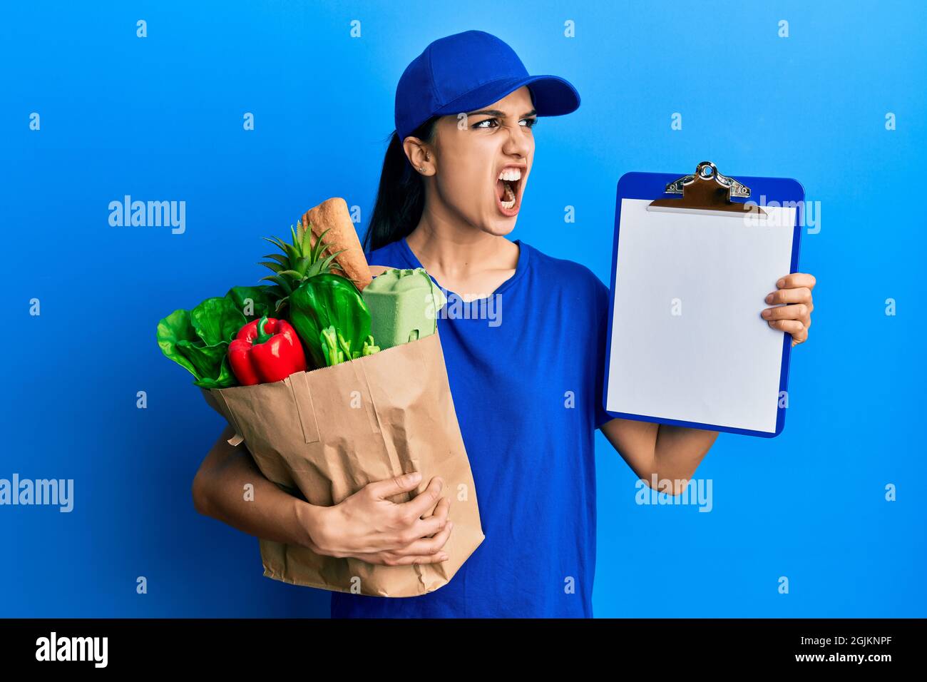 Young hispanic woman wearing courier uniform with groceries from supermarket and clipboard angry and mad screaming frustrated and furious, shouting wi Stock Photo