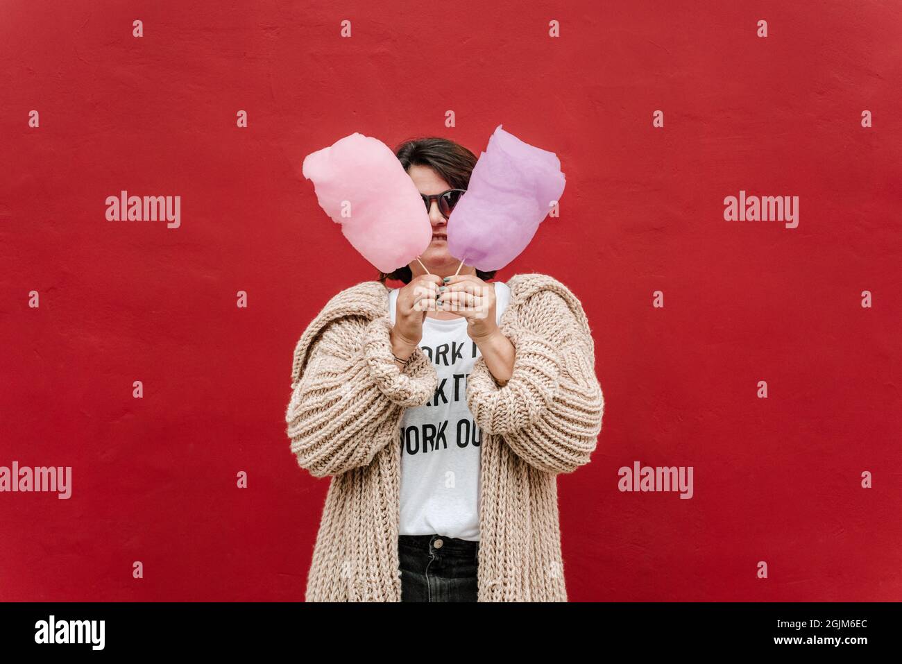 image of a woman with two cottons of sugar, pink and lilac. She wears dark glasses, bundled up with a sweater on a red background. Copy space Stock Photo