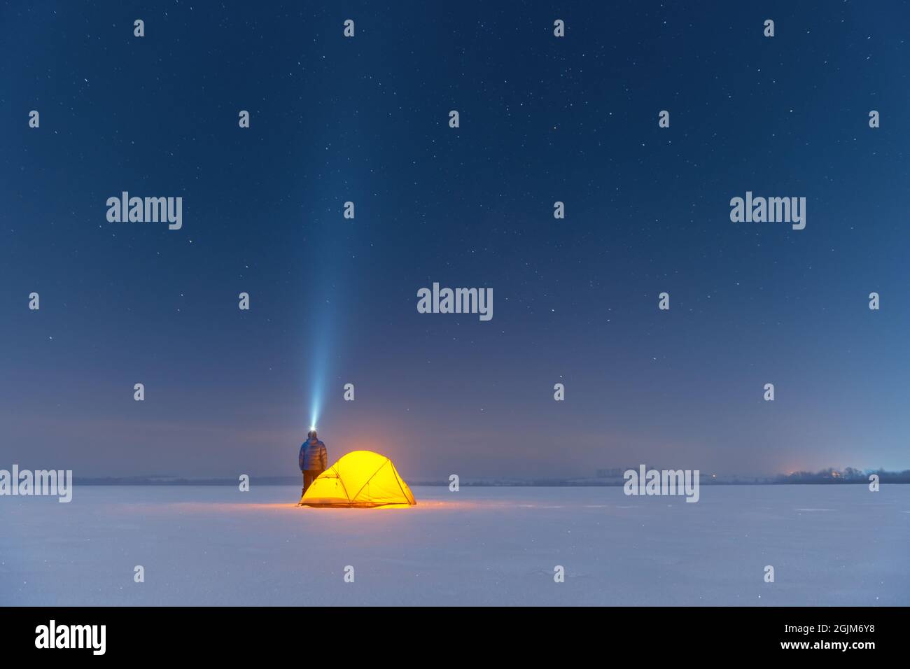Tourist with flashlight near yellow tent lighted from the inside against the backdrop of incredible starry sky. Amazing night landscape. Tourists camp in snowy field. Travel concept Stock Photo