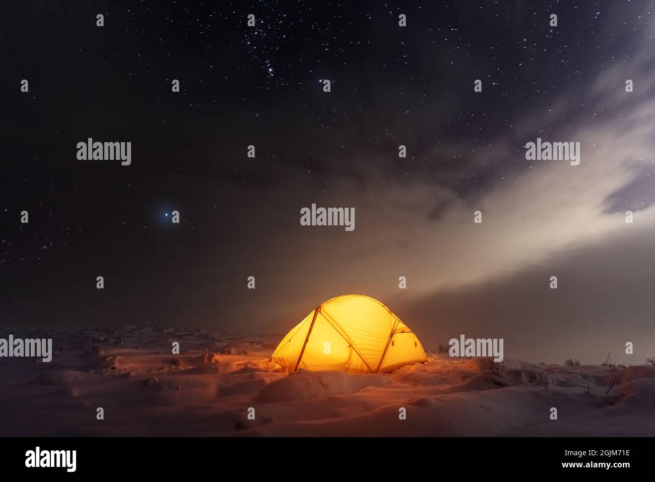 Yellow tent lighted from the inside against the backdrop of glowing city lights and incredible starry sky. Amazing night landscape. Tourists camp in winter mountains. Travel concept Stock Photo