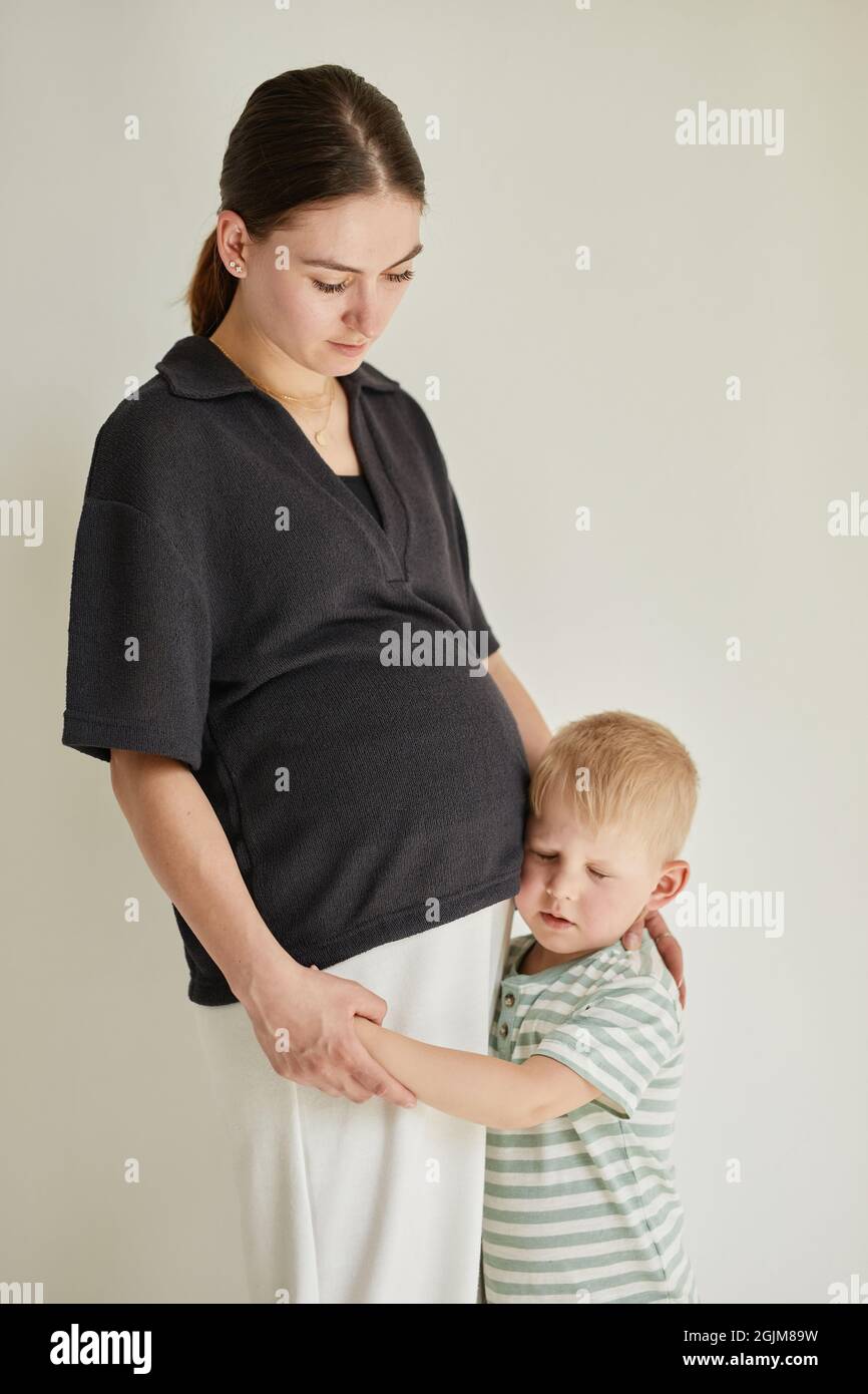 Blond-haired son in stripped tshirt focused on heartbeat of baby in mothers belly while keeping ear near it Stock Photo