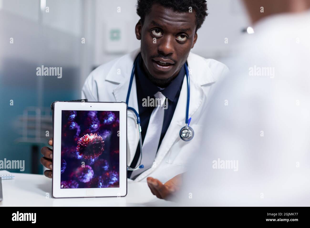 Doctor of african american ethnicity holding digital tablet with virus animation while sitting at desk. Black medic explaining disease bacteria and healthcare problems to old patient Stock Photo