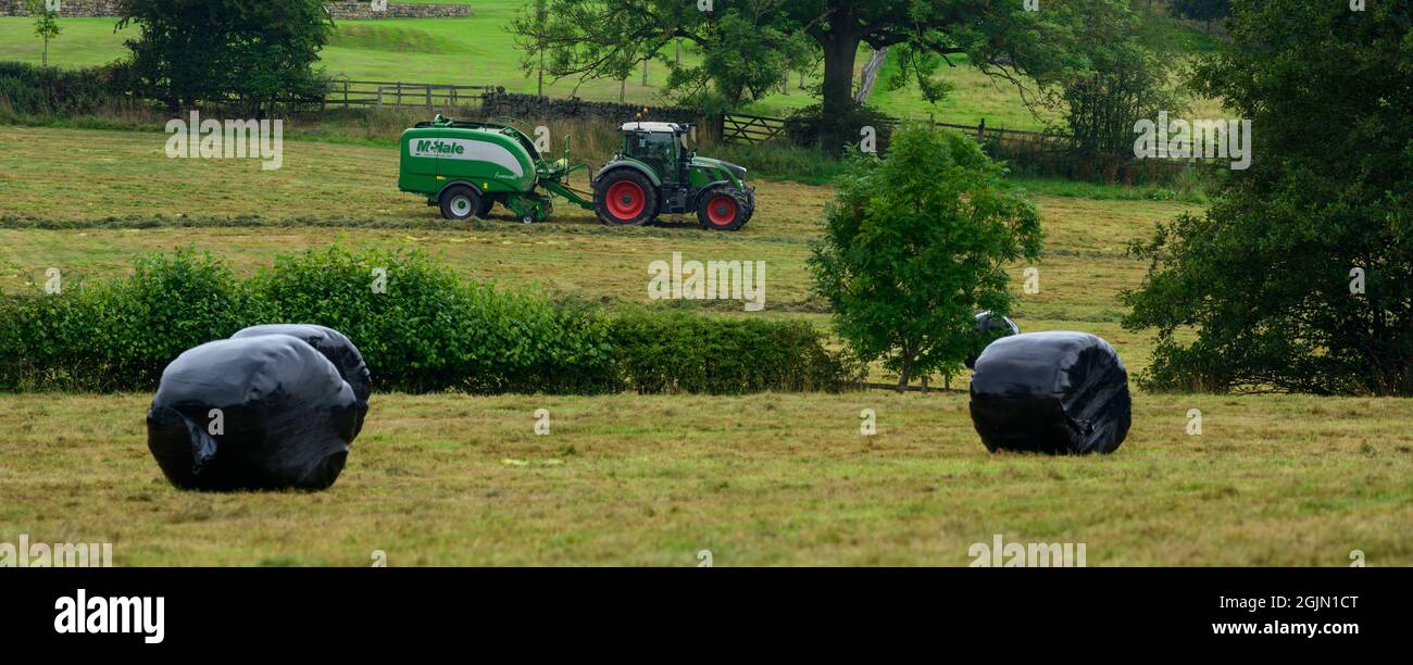 Hay & silage making (farmer in farm tractor at work in rural field pulling baler, collecting wrapping dry grass in round bales) - Yorkshire England UK Stock Photo