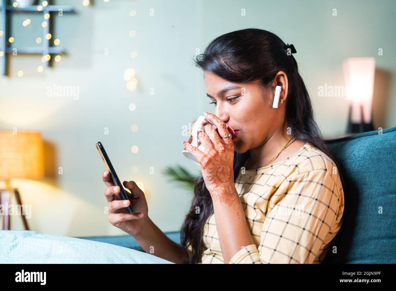 Happy smiling young girl busy using social media or chatting on mobile phone by having coffee while sitting on sofa - concept of millennial using Stock Photo