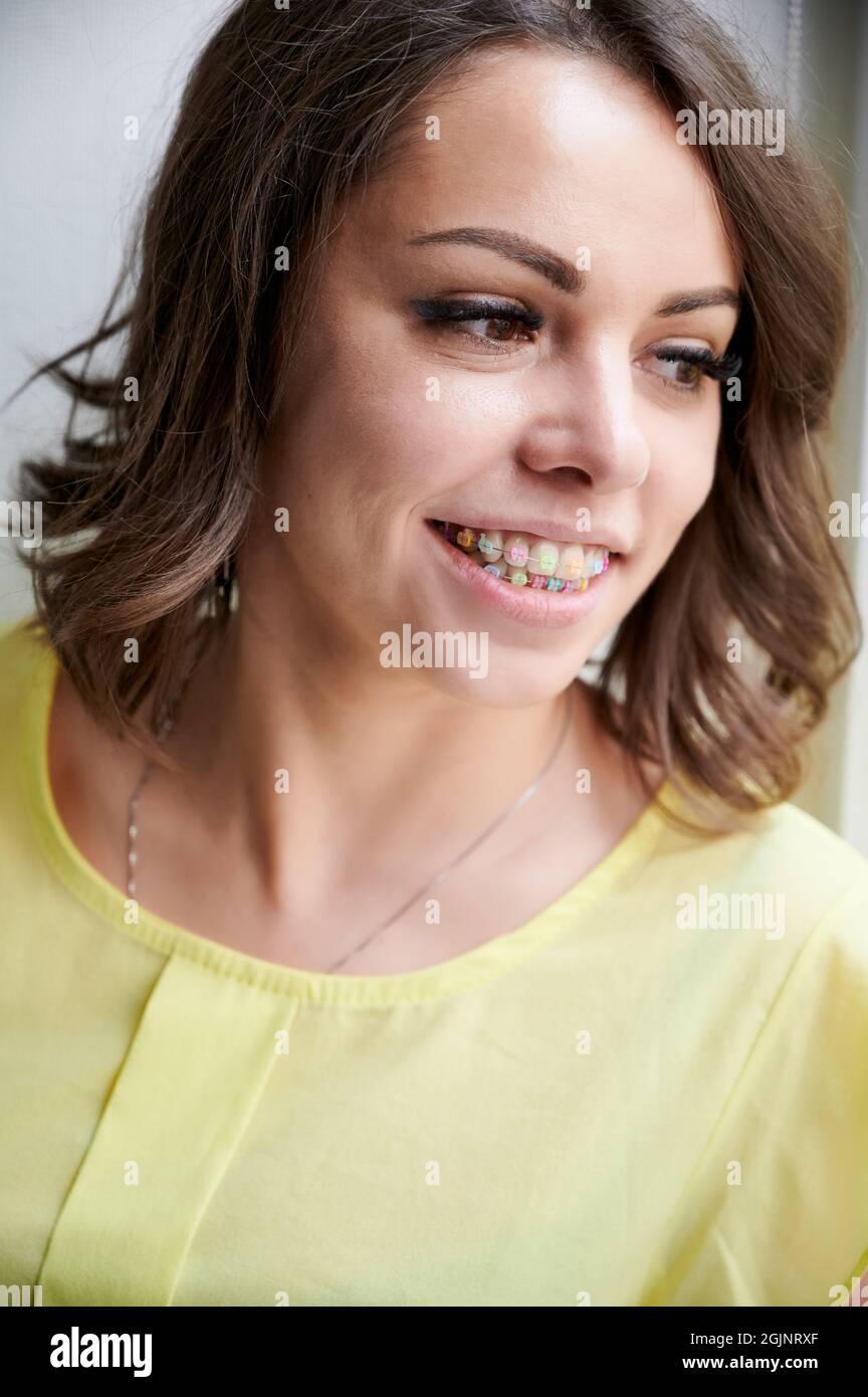Portrait of cheerful woman with orthodontic brackets. Joyful woman with brown hair showing braces with multicolored rubber bands on her teeth. Concept of stomatology and orthodontic treatment. Stock Photo