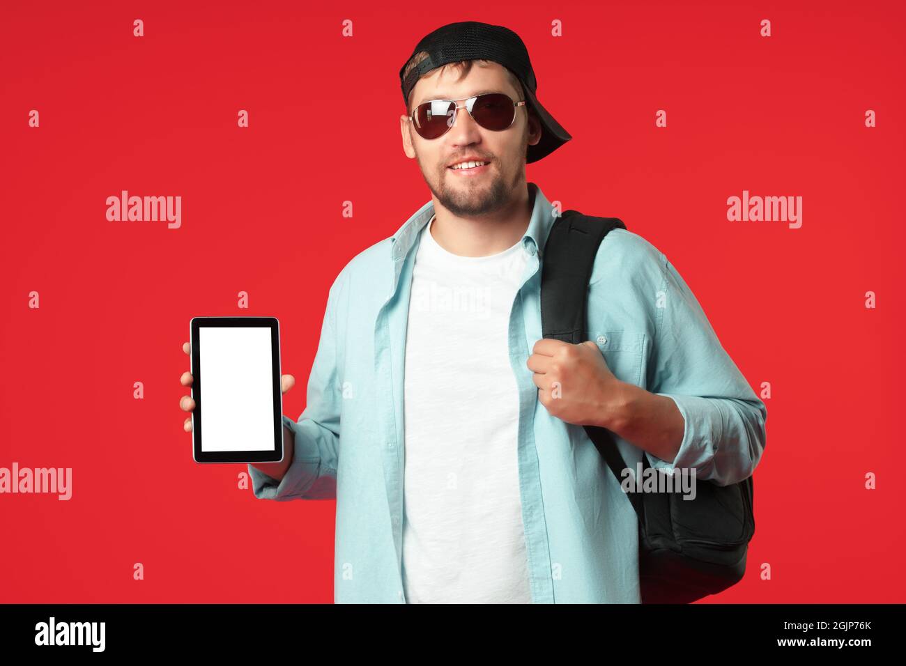 A European tourist in a baseball cap and sunglasses with a backpack on his shoulder stands on a red background and shows a tablet. The concept of a mobile application for travelers. Mockup Stock Photo