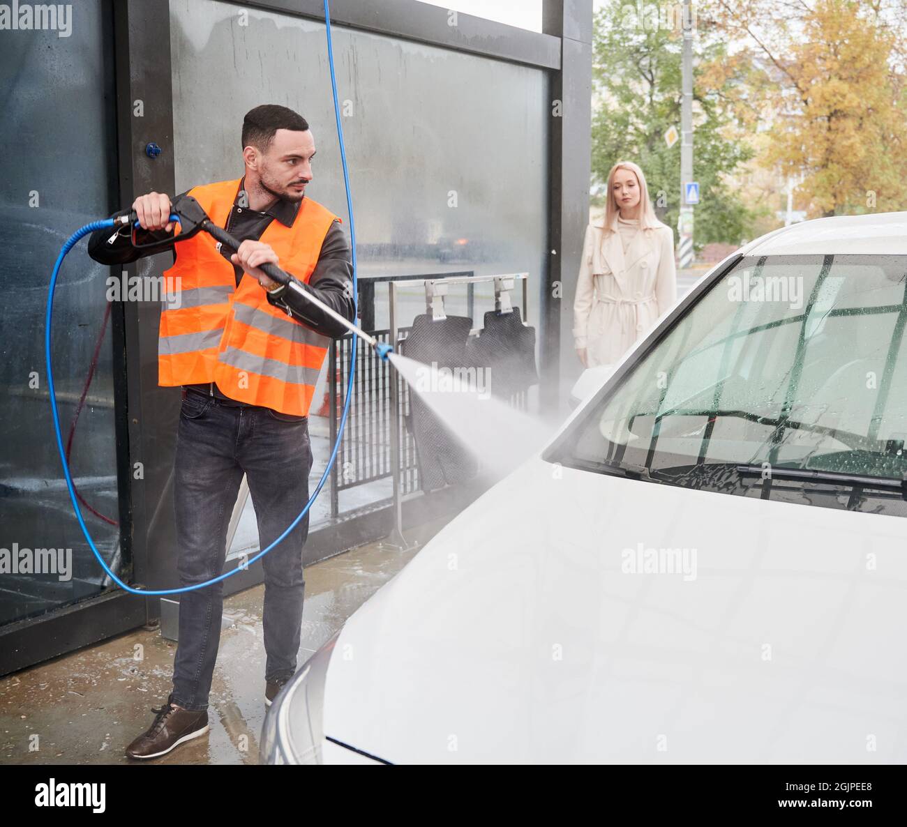 Young man washing car on carwash station outdoor, wearing orange vest. Handsome worker cleaning automobile, using high pressure water. Stock Photo