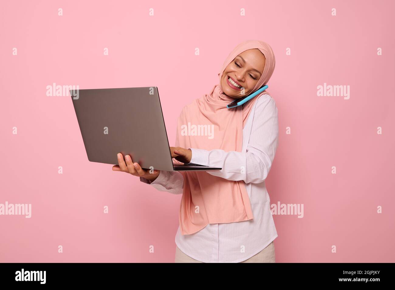Successful Muslim Arab gorgeous woman in hijab, dressed business smart casual talks on mobile phone , smiles looking at the monitor screen while typin Stock Photo