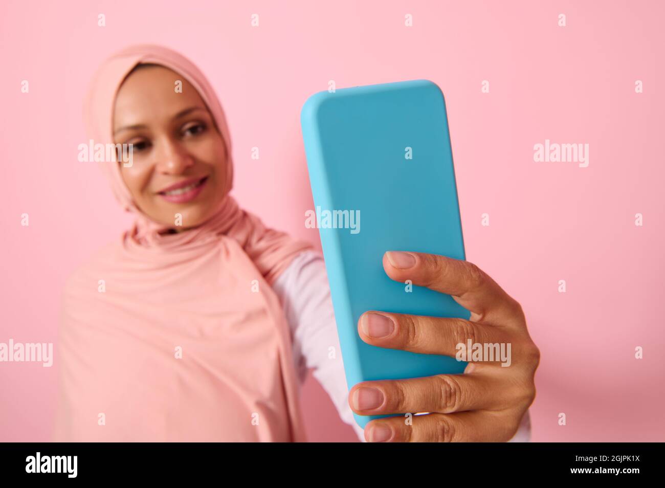Soft focus on the smartphone in blue cover in outstretched arms of Arab Muslim woman wearing traditional religious islamic outfit, pink hijab,and smil Stock Photo