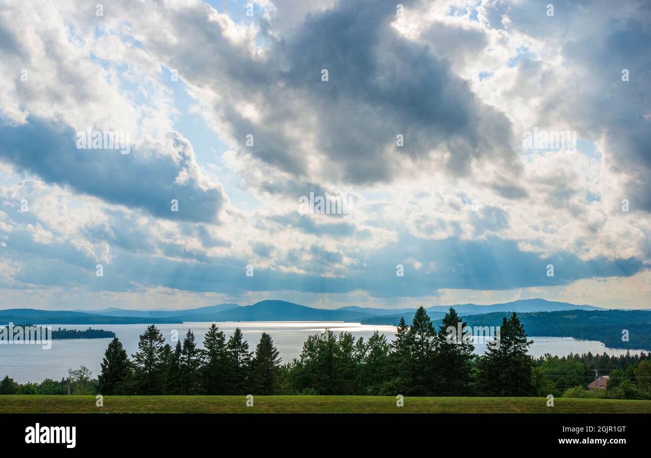 Scenic view of the Rangeley Lake, the Bald Mountain and distant ridges of the Boundary Mountains, behind a spruce grove, under cloudy skies. Stock Photo