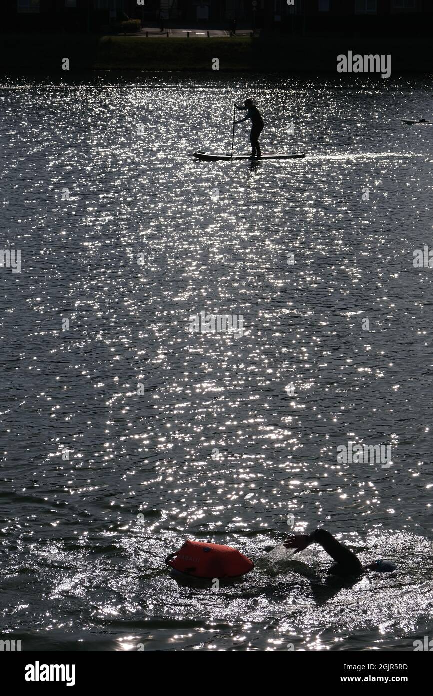 An open water swimmer with safety float in Llanelli's North Dock (now a leisure facility) with a paddle boarder silhouetted in the background. Stock Photo