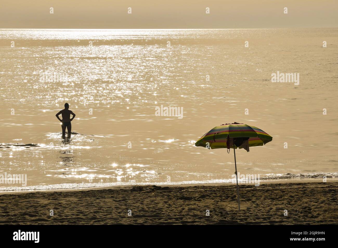 Rear view of the silhouette of a man standing in the seashore at sunset with a sun umbrella on the sandy beach, Marina di Castagneto Carducci, Tuscany Stock Photo