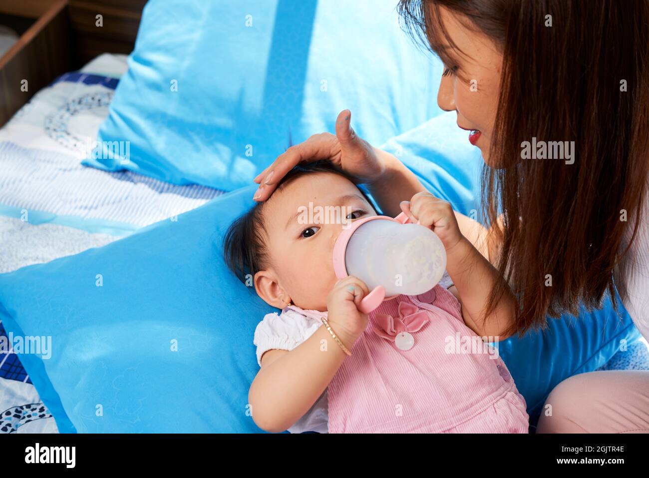 Baby girl drinking formula with vitamins and looking at camera when mother is stroking her head softly Stock Photo