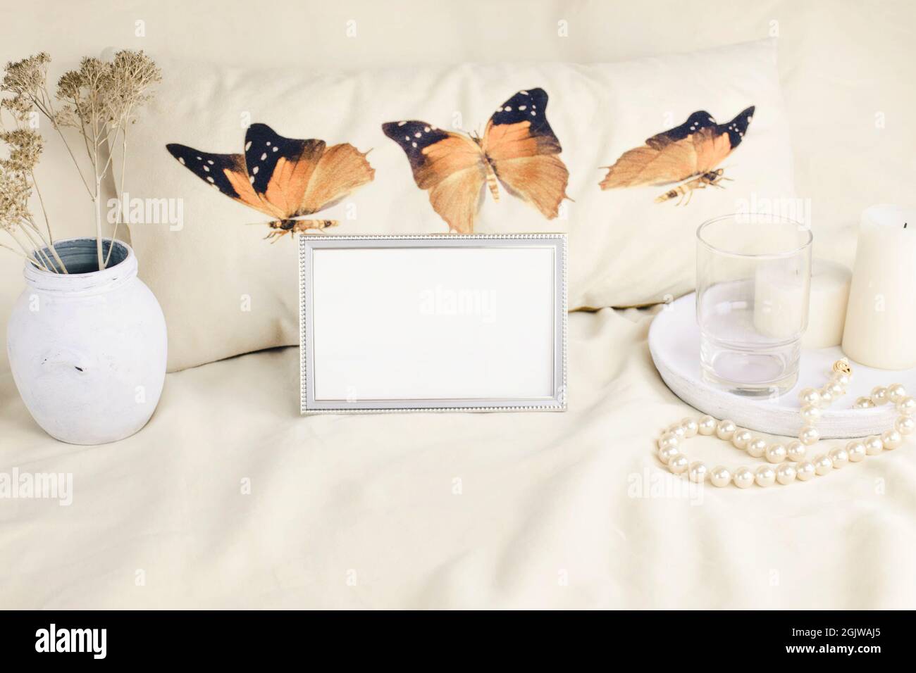 Portrait metal frame mockup on the background of a pillow with butterflies, a vase, white candles and a necklace of pearls, an empty glass on a white Stock Photo