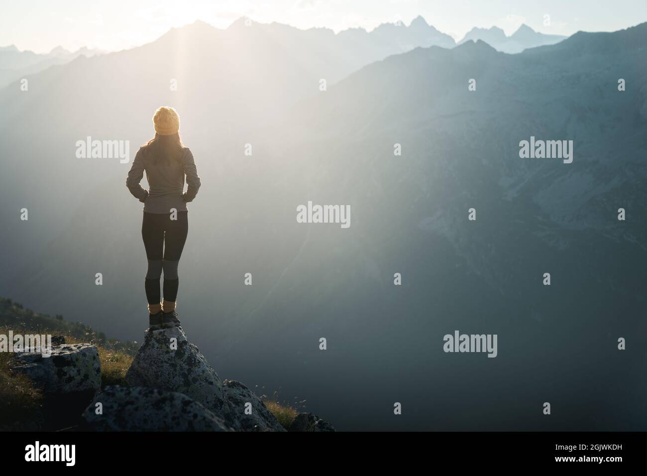 Young woman hiker standing on the edge of cliff at sunrise. Female tourist reaching summit enjoying amazing sunrise in the mountains.Backlight sunligh Stock Photo