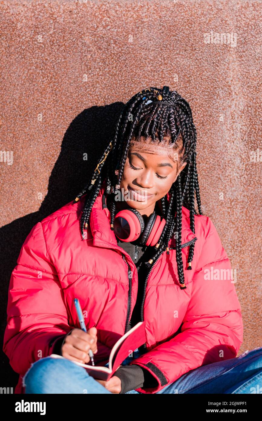 Beautiful African woman wearing red jacket with headphones outdoors. Black woman writing in a notebook Stock Photo