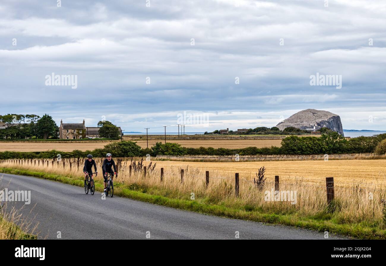 Two male cyclists cycling on country road with Bass Rock gannet colony in the distance, East Lothian, Scotland, UK Stock Photo
