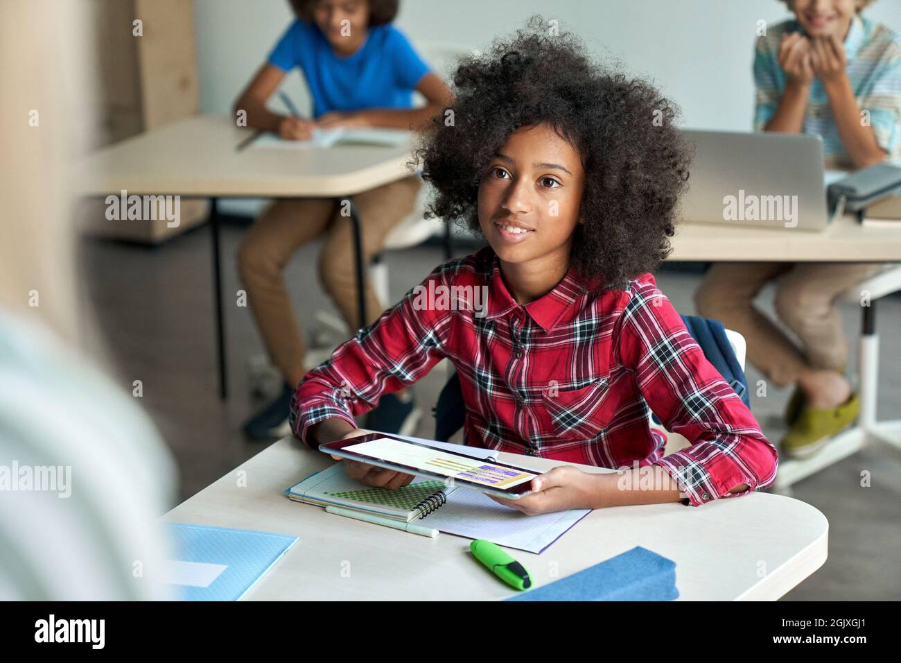 Happy Afro American schoolgirl looking at teacher holding tablet in classroom. Stock Photo