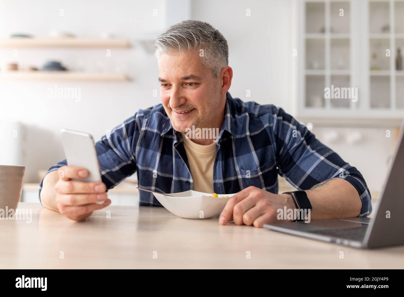Happy mature man using smartphone while having breakfast in kitchen table at home, copy space Stock Photo