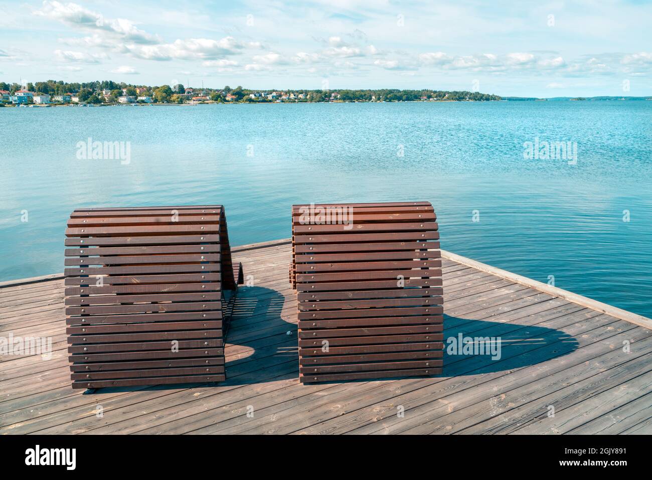 Two wooden beach beds or chairs on a wooden pier next to the sea with cityscape in the background. Sunny summer day by the Baltic Sea in Vastervik Stock Photo