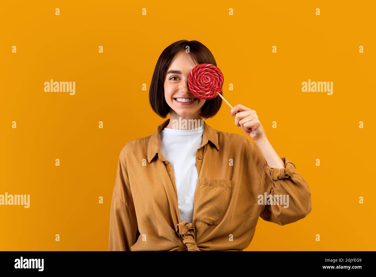 Sweet tooth concept. Happy young lady holding candy on stick, covering one eye with lollipop, yellow studio background Stock Photo