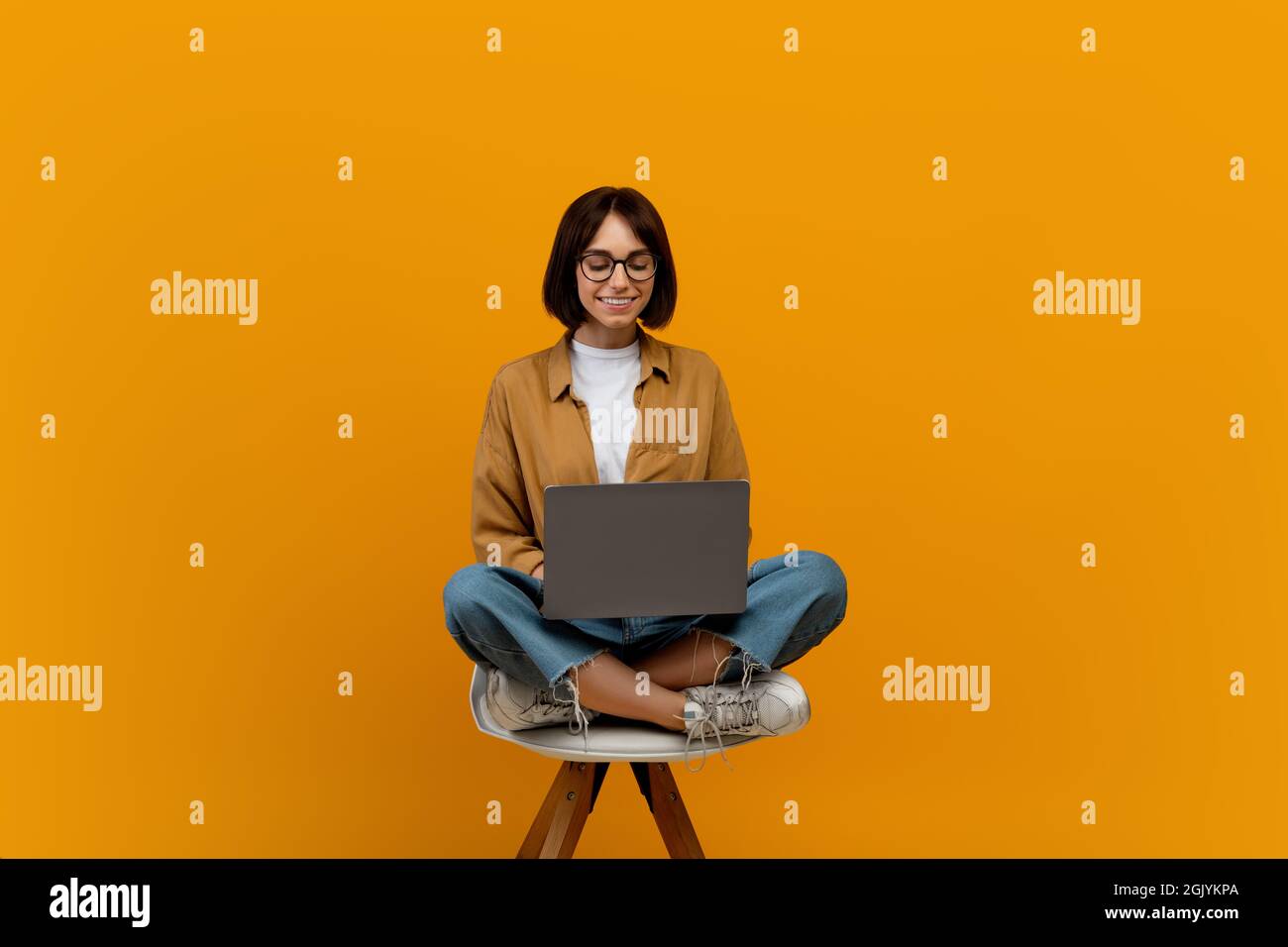 E-learning concept. Female student using laptop computer while sitting in chair over yellow background, copy space Stock Photo