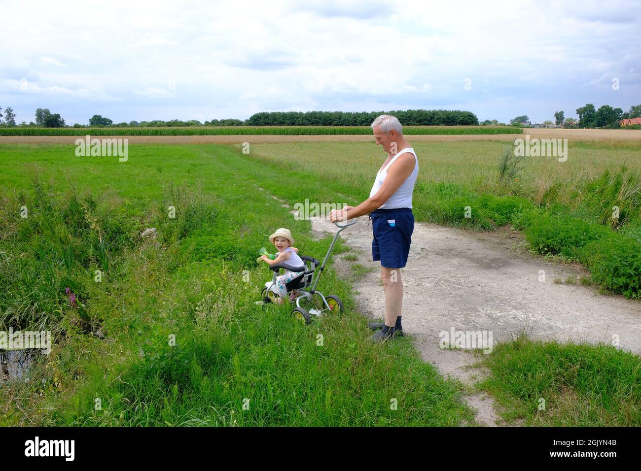 Full-length view of grandpa and granddaughter. A smiling little girl sits on a bicycle in the meadow. Senior man is standing behind girl Stock Photo