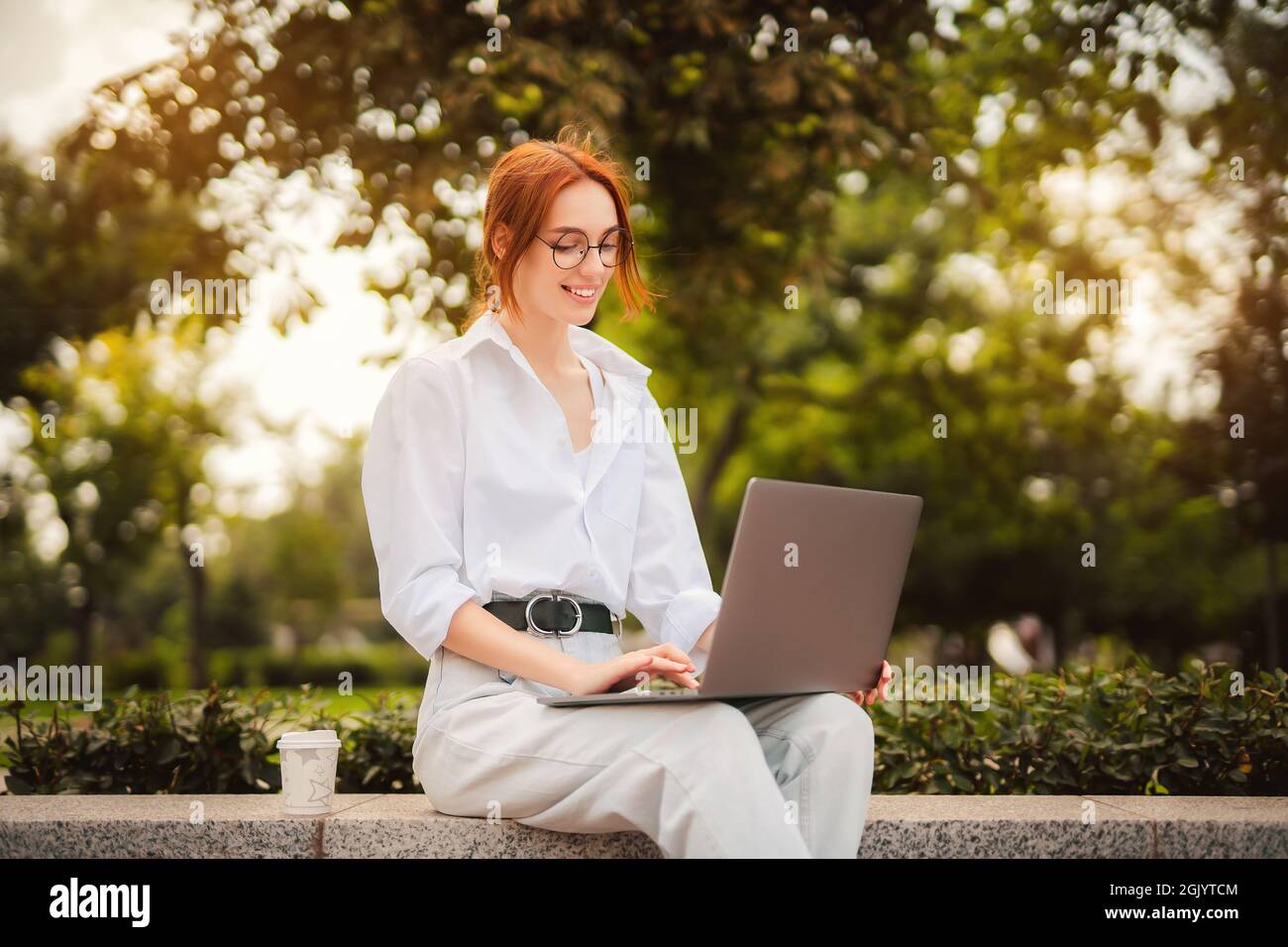 Beautiful redhead young woman sitting in the park and using laptop ...